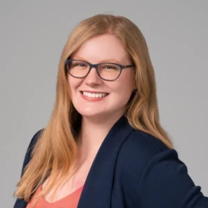 Kayla Adamek headshot. She has long strawberry blonde hair and glasses. She is wearing a blue blazer with a pink shirt under it.