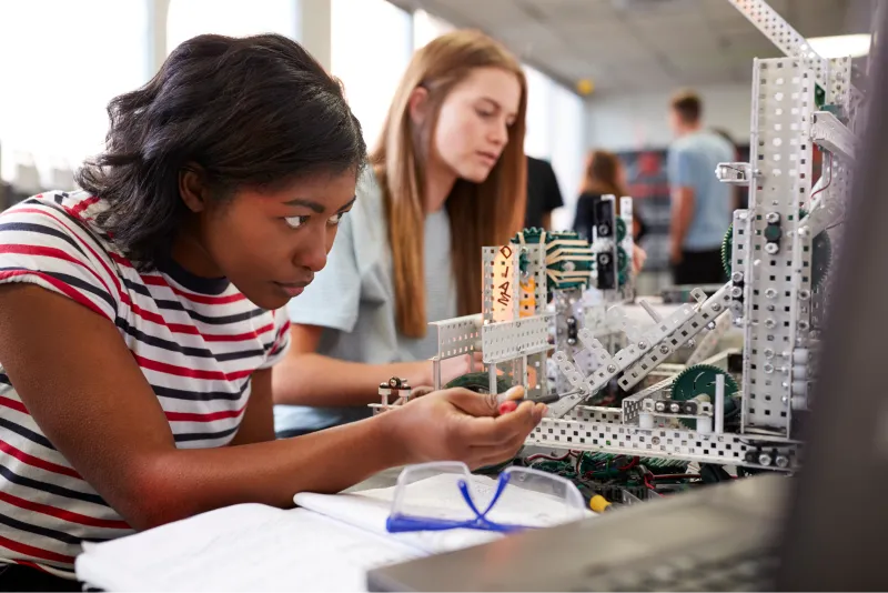 Two Female College Students Building Machine In Science Robotics Or Engineering Class.