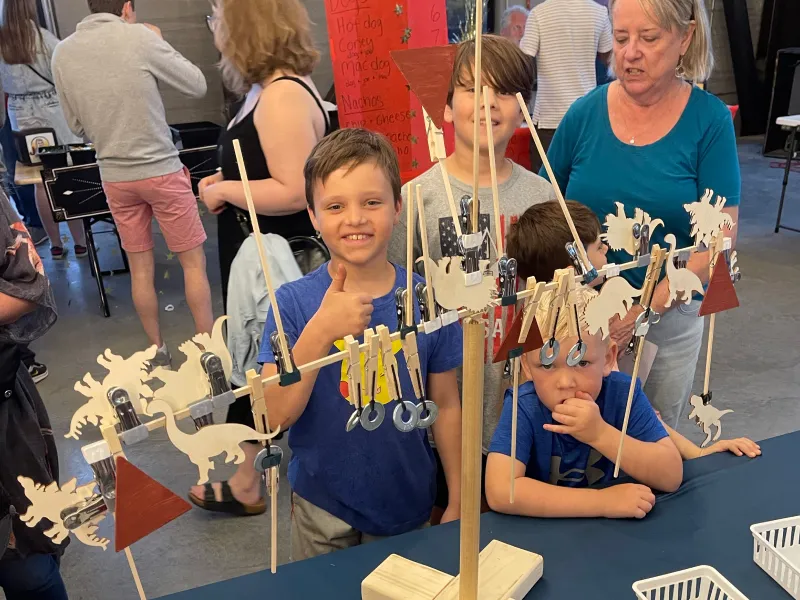 A young boy giving a thumbs up and smiling with pride as he utilized all the items that could be hung on the wooden dowel to make both sides balance. It is covered in metal washers and small wood dinosaur cutouts.