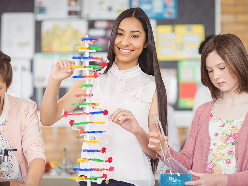 Girl with long straight black hair engaging with a DNA model activity. Girl next to her holding a beaker with blue liquid in it